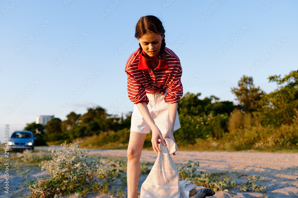 Obraz premium Woman in a striped red top and white shorts bends to pick up litter on a sandy beach, bag in hand, under a clear blue sky. Outdoor lifestyle shot highlighting environmental responsibility.