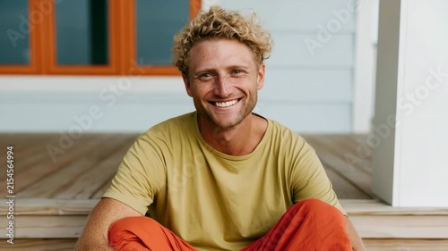 Seated young man with curly hair in mustard shirt and orange pants on wooden porch for relaxed portrait background