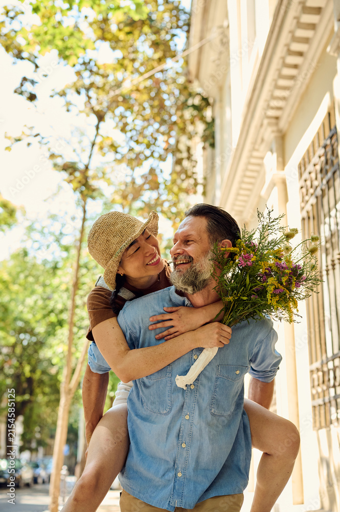 Fototapeta premium Happy Couple Piggyback On Urban Street With Bouquet — Asian Woman And Bearded Man Laughing