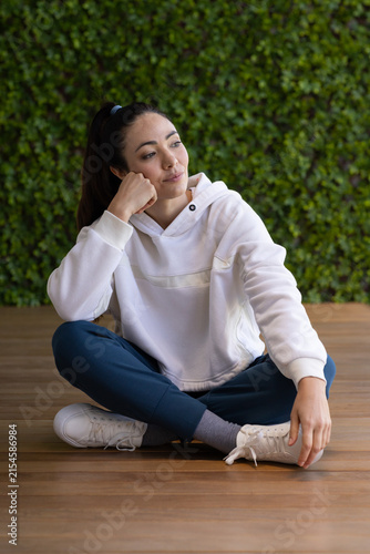 Asian adult female sitting cross-legged on wood deck by green plant wall, touching untied sneaker