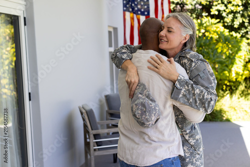 Diverse couple embracing on front porch, woman in camouflage holding patrol cap near American flag
