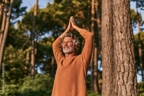 A silver-haired man in a ribbed orange sweater practices mindful yoga with hands joined overhead surrounded by a serene sunlit forest grove at dusk