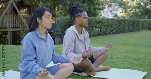 Diverse friends sitting on yoga mats on lawn after meditating lifting arms overhead for centering