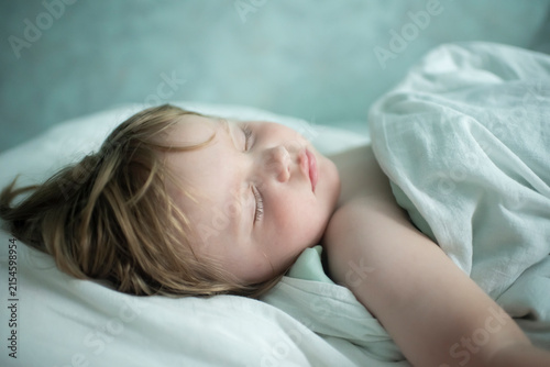 Cute toddler boy sleeping peacefully in bed under blanket at home