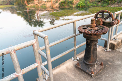 Rusty metal gear mechanism by a calm water canal, surrounded by a natural outdoor setting, showing industrial decay.