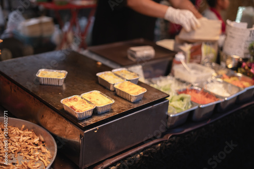 Baked food trays on a grill at a street food market, with fresh vegetables on the side.