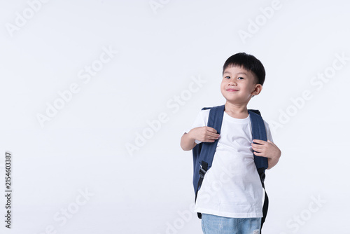 Confident young boy with a playful smirk, wearing a white t-shirt, jeans, and a backpack, posing in a bright studio.