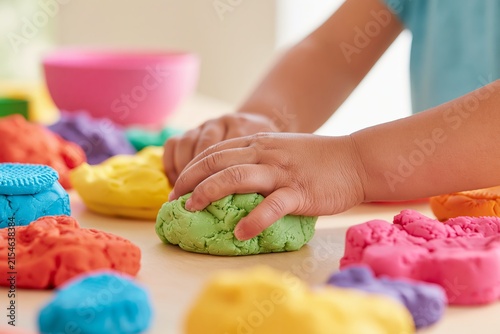 Close up of child's hands squishing colorful playdough on a wooden table with various vibrant playdough shapes and a pink bowl in the background