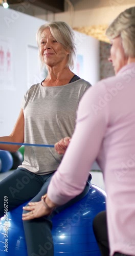 Vertical video: Female senior and trainer touching shoulder and thigh pulling band on ball in rehab