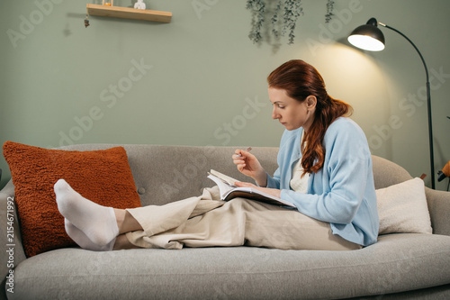 Redhead woman relaxing on cozy sofa and writing notes in notebook at home during evening study or personal planning session