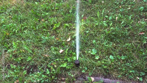 Close view of a small ground sprinkler spraying a continuous stream of water upward over green grass and weeds in an outdoor garden or yard