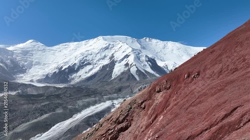 Aerial video pan revealing snow-capped Lenin Peak behind a steep red mountain slope under a clear blue sky