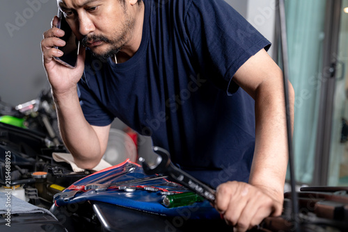 Young man standing near his broken down car, checking the open engine hood while making a phone call for roadside assistance.