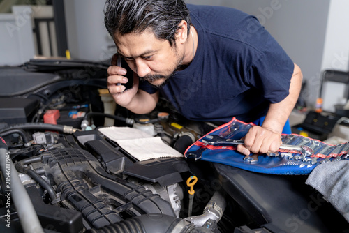 Young man standing near his broken down car, checking the open engine hood while making a phone call for roadside assistance.