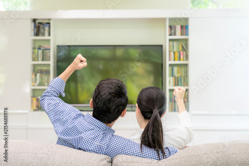 A young couple watches a sports game on television and cheers together in their living room at home.