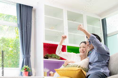 A young couple watches a sports game on television and cheers together in their living room at home.