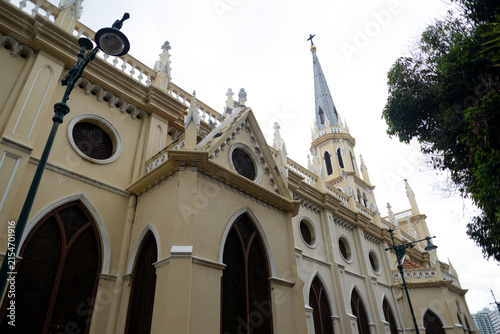 Elegant Gothic Revival church facade featuring pointed arches, ornate stone details, circular windows, and a tall central spire against an overcast sky.