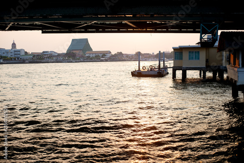 Scenic view under a steel bridge over a wide Chao Phraya river during sunset with golden light shimmering on water ripples at dusk with traditional riverside architecture and a temple silhouette.