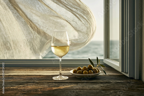 Glass of chilled white wine with a bowl of olives by a sea-view window