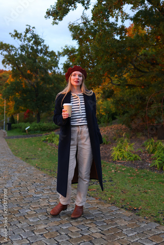 Stylish young woman walking in autumn park with takeaway coffee, wearing a beret and long coat.