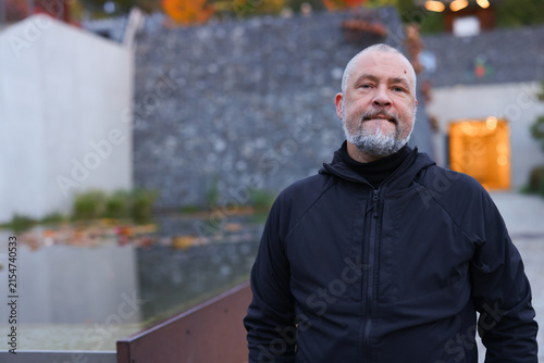 Mature bearded man in black jacket standing outdoors in a modern urban setting, confident male lifestyle portrait.