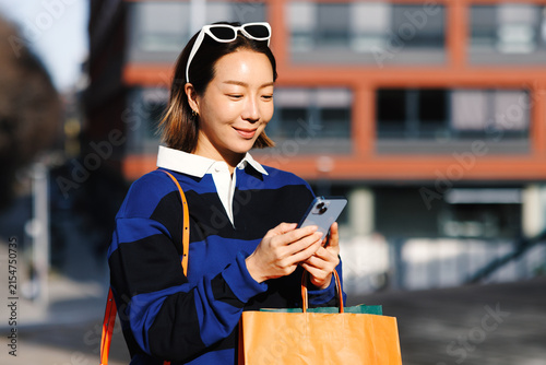 Foto Young asian woman smiling looking at her smartphone and holding shopping bags in