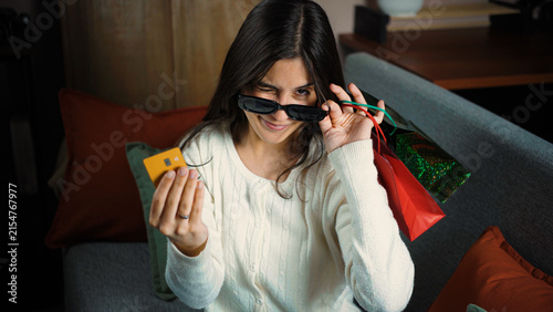 Fotografie Young Latina woman winking while sitting on her sofa and holding shopping bags and a credit card, conveying a playful and satisfied shopping mood