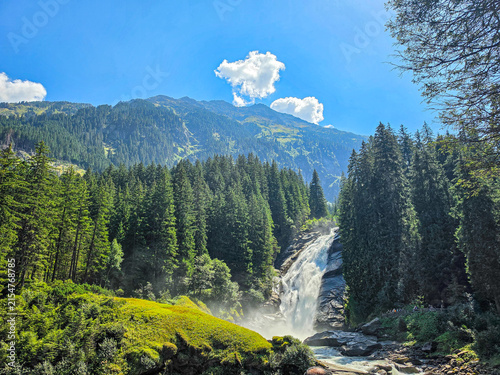 powerful alpine waterfall cascading through lush evergreen forest beneath a dramatic mountain landscape