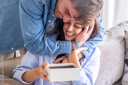 Fotografie Man hugging smiling woman as she opens a gift box at home, romantic surprise bet