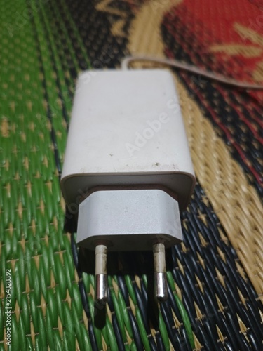 Foto Foto menampilkan sebuah adaptor atau kepala charger berwarna putih yang diletakkan di atas kain tenun bermotif warna-warni
