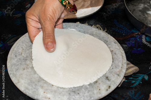 Photography Hand lifting a freshly prepared round rice flour roti from a traditional round marble chakla or rolling board, showing a step in the preparation of homemade Indian rice flour flatbread