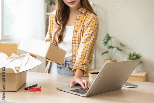 Online sell store, startup small business entrepreneur asian young woman using laptop computer checking purchase product order from web, owner working at home office, prepare pack parcel for delivery.