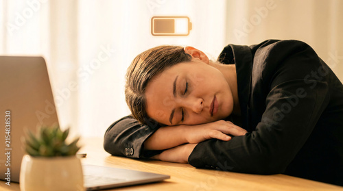 Woman sleeping at desk, head resting on arms, low battery icon above head, symbolizing burnout and exhaustion, workplace fatigue, overwork concept
