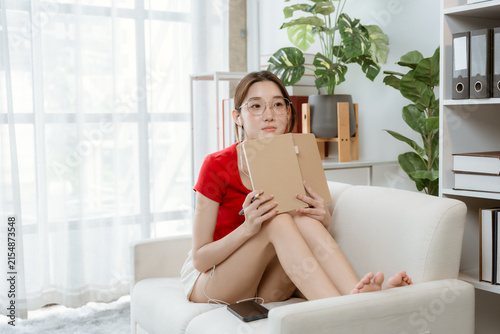 Canvas Print A Young Woman Lost in Thought: A young woman in a red top and shorts sits on a comfortable sofa, holding a book, with a pensive expression as she gazes into the distance
