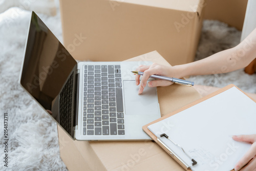 Canvas Print Unpacking and Organizing: A close-up shot of hands writing on a clipboard next to an open laptop, surrounded by cardboard boxes, suggesting moving or organizing items