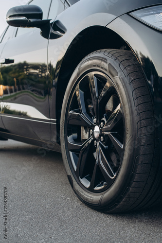 Close up of a black car wheel showing the glossy alloy rim, detailed tire tread, curved bodywork, and side mirror on a paved outdoor surface.