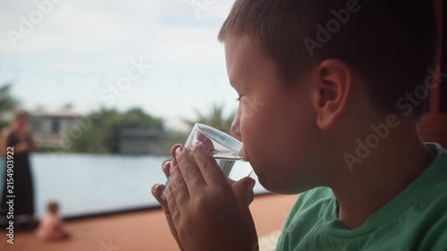 Young autistic child drinking water from a glass by a swimming pool, enjoying a calm outdoor moment with family during a summer day.