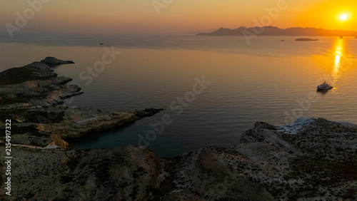 Golden sunlight rises over the rocky coastline of Mandrakia on Milos, reflecting across calm water and illuminating the rugged cliffs of the Aegean in early morning light.