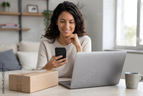 Plano medio de mujer sonriente mirando su teléfono inteligente junto a una caja de cartón y ordenador portátil en escritorio de salón con iluminación natural.