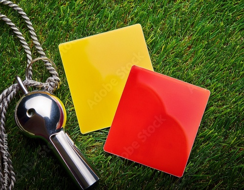 bright yellow and red penalty cards with a silver referee whistle lying on the green grass of a soccer field