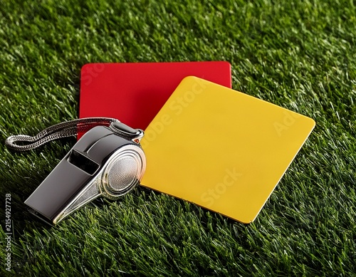metal referee whistle and bright yellow and red penalty cards resting on the green grass of a soccer field pitch