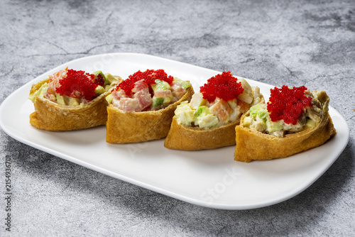 Delicious Japanese inarizushi stuffed with salmon, avocado, and topped with flying fish roe.  Inari sushi set with seafood salad, avocado, and red tobiko caviar served on a white plate.
