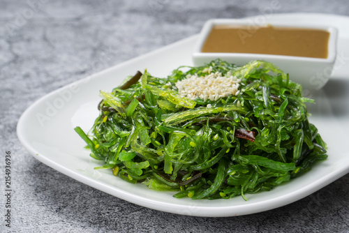 Fresh hiyashi wakame salad with sesame seeds and peanut dressing against grey concrete background. Green seaweed chuka salad on a white plate with sauce bowl, close up view with copy space.