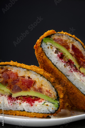 Crispy tempura sushi burger cut in half showing layers of tuna tartar, avocado, cream cheese and red tobiko. Crunchy fusion sushi hot dog burger close up with fresh seafood and rice inside against dar