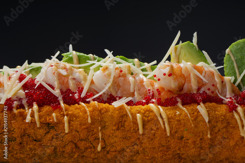 Corn dog sushi with shrimp, avocado, sprinkled with Parmesan cheese, and drizzled with sauce. On a black background.