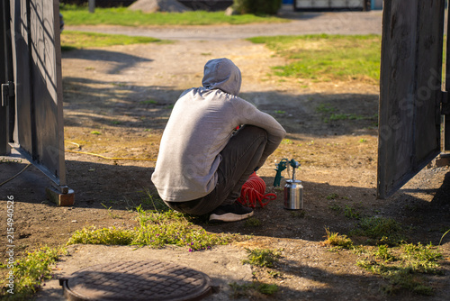 Fotografía Worker wearing hoodie sitting on haunches next to pneumatic spray gun on ground outdoors between open metal gate doors during break or preparation