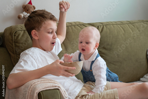 Fotografía Older brother shares snack with baby sibling while relaxing together on sofa at home