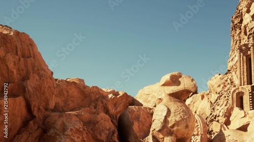 A large stone figure stands in a desert area. The landscape shows rocks and ancient ruins in the background. The sun shines brightly in a clear sky above the scene.