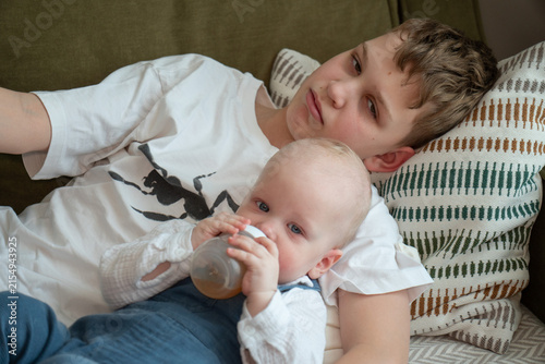 Fotografía Older brother cuddles baby sibling while younger child drinks from bottle resting safely in his arms on sofa at home