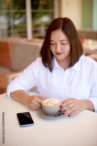 woman with cup of cappuccino and mute phone sitting at outdoor cafe